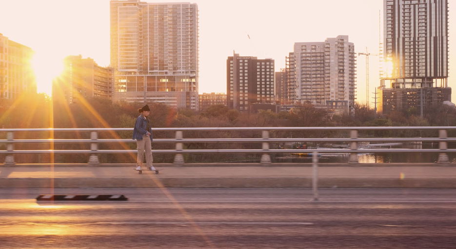 Young woman skateboarding on sidewalk with sun setting behind city in background
