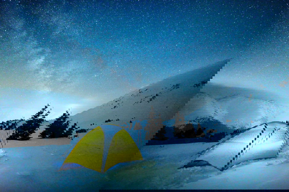 Illuminated tent pitched in snowy mountains at night beneath starry sky