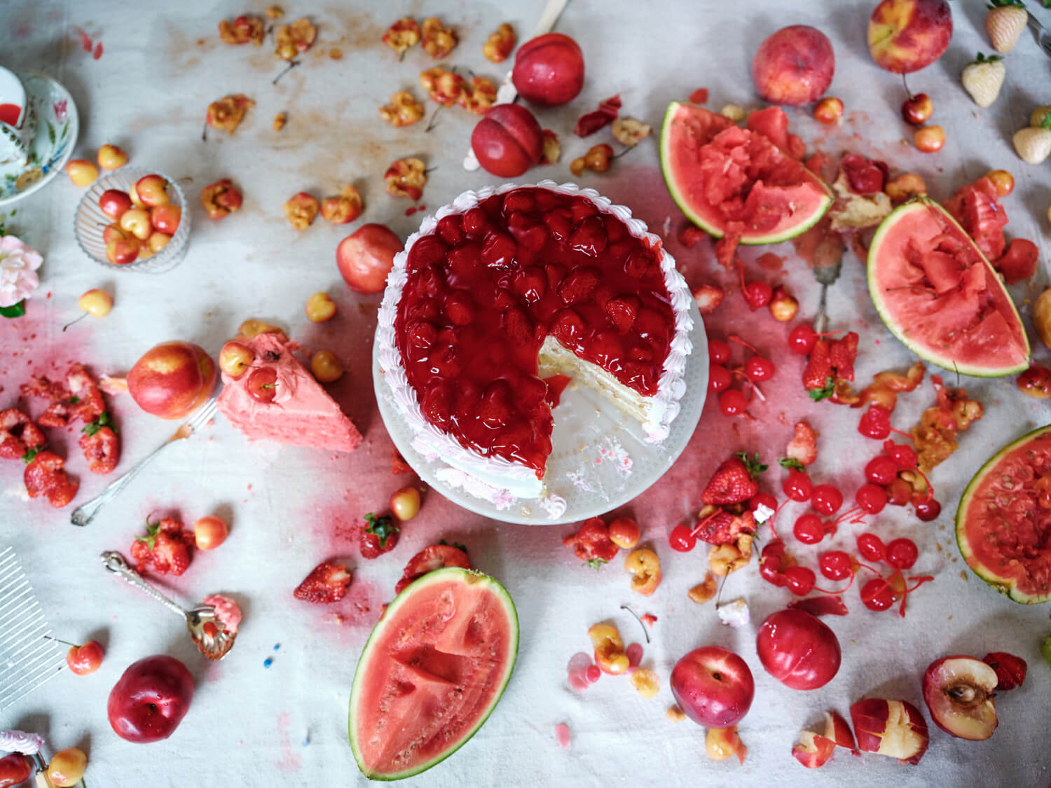 Flat lay of cake covered in strawberries on a cake dish upon a table strewn with squashed fruit that has stained the table cloth red.