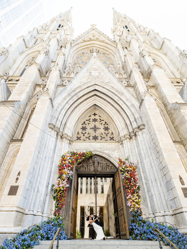 Wide angle photograph of bride and groom kissing on the steps of a cathedral with towers stretching above them in converging verticals