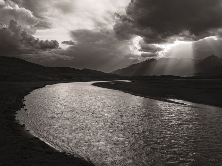 Moody black and white landscape photograph of meandering river banked by mountains and lit by rays of sunshine streaming through a break in dark clouds