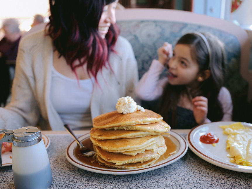 Pancake stack on diner table in front of woman and young girl who are looking st each other smiling in excitement