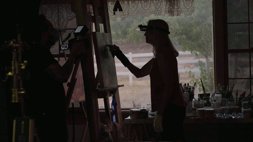 Videographer, Giulio Meliani uses FUJINON XF50mmF1.0 R WR to profile legally-blind artist, Therese Verner and show the importance of art. Image shows video still of Therese Verner painting on an easel in front of a window in her art studio.