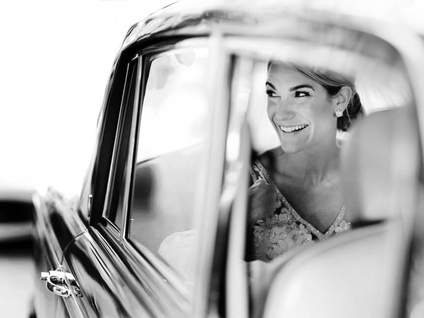 Professional photographer, Alison Conklin, on why the FUJIFILM GFX system is so good for location portrait photography. Black and white image through the window of a classic car of a smiling bride who is sitting in the back seat.