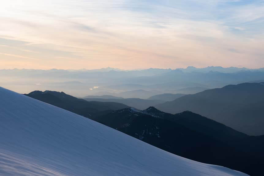 A snowy slope in the foreground. A background of stretching mountain peaks and valleys, foggy and indistinct