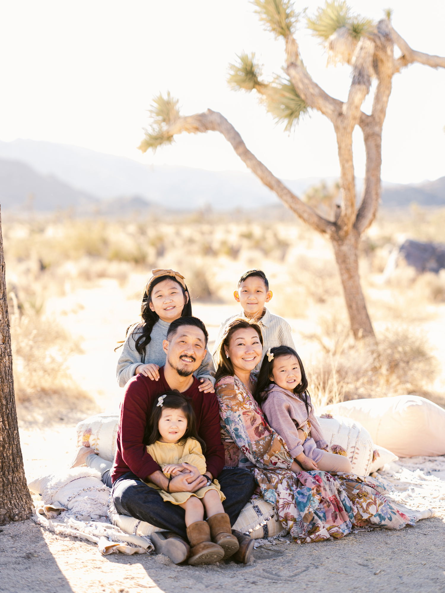 An Asian family of six huddle together for a portrait, amongst sand and trees. Their smiles are genuine and sincere
