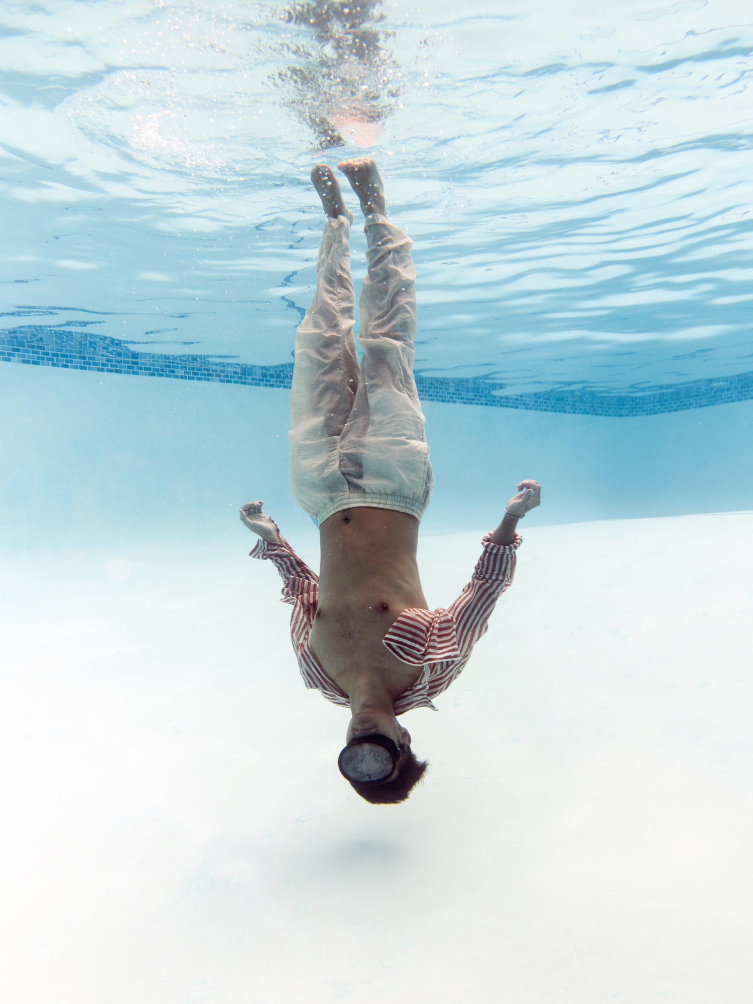 Dressed in a red pinstripe shirt and white trousers, a man poses upside-down underwater