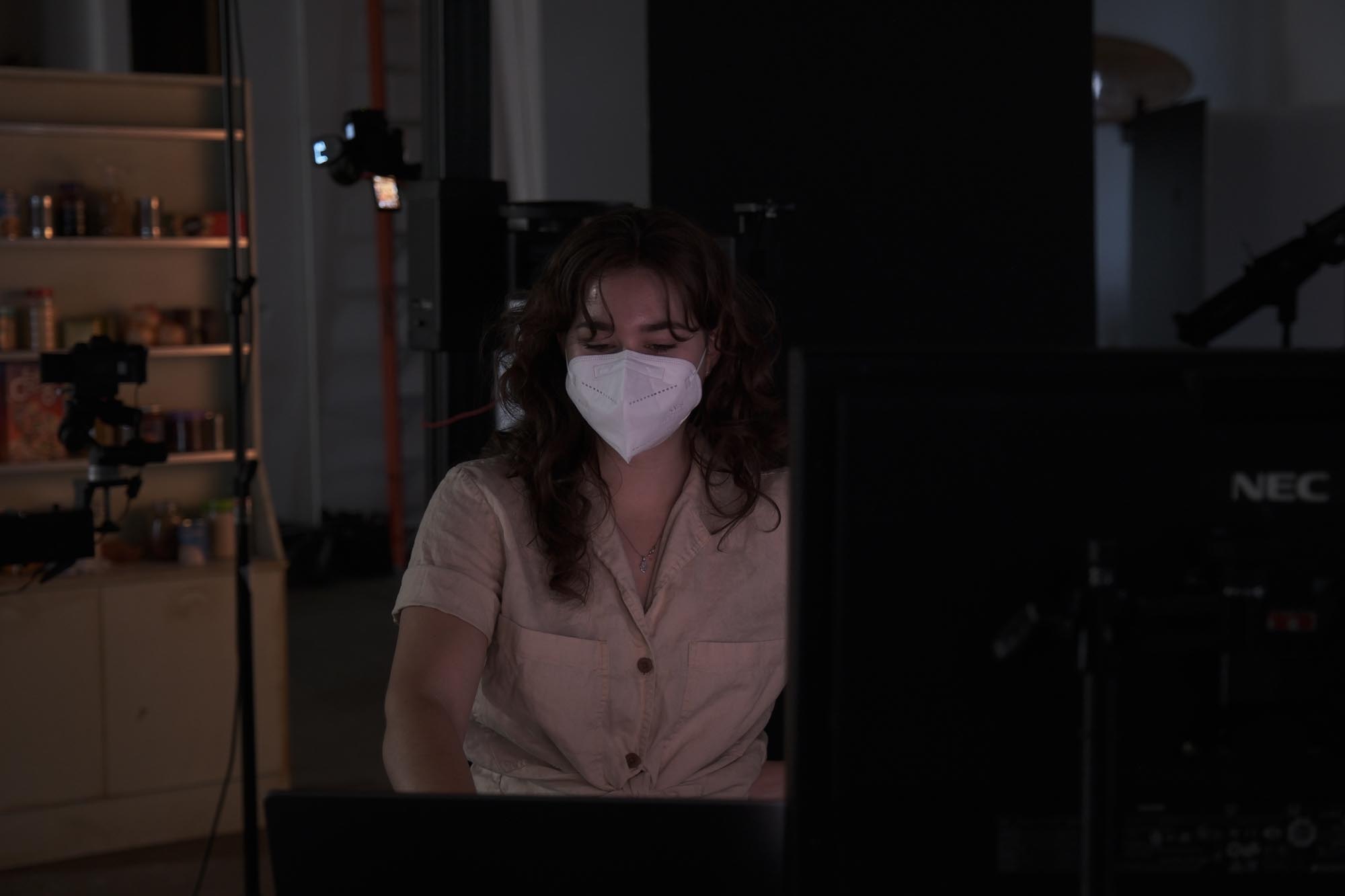 A masked woman surveys a studio setup, monitoring a food commercial shoot