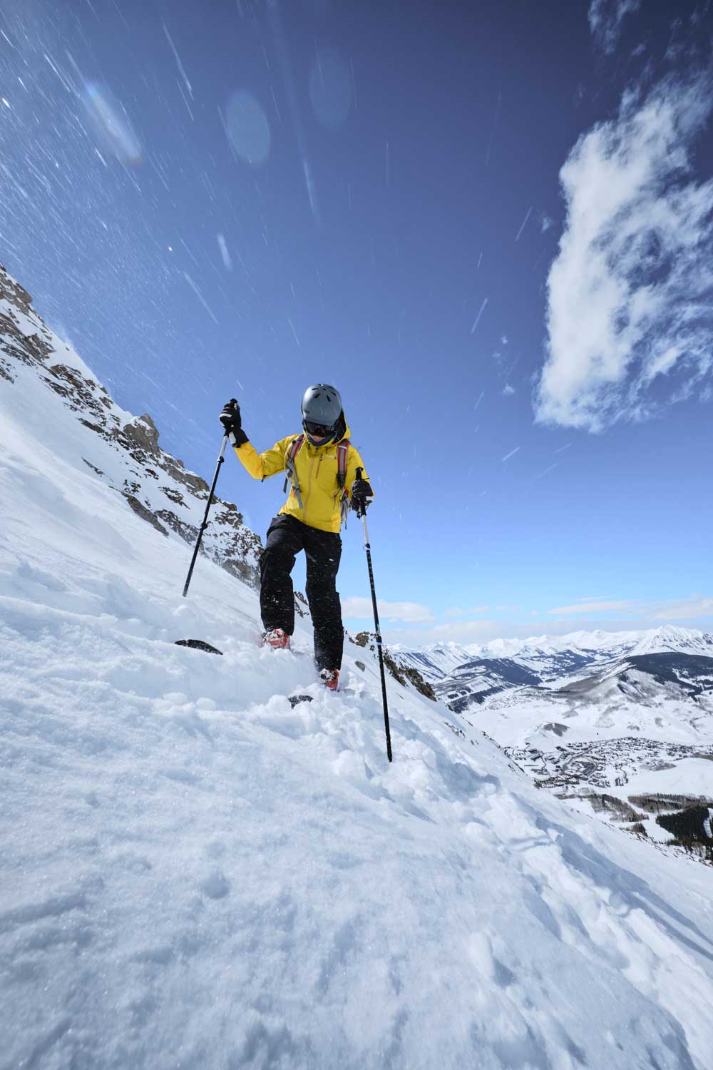 Wide-angle image from low angle showing a man on skis wearing yellow jacket traversing a steep slope with ski poles in hand