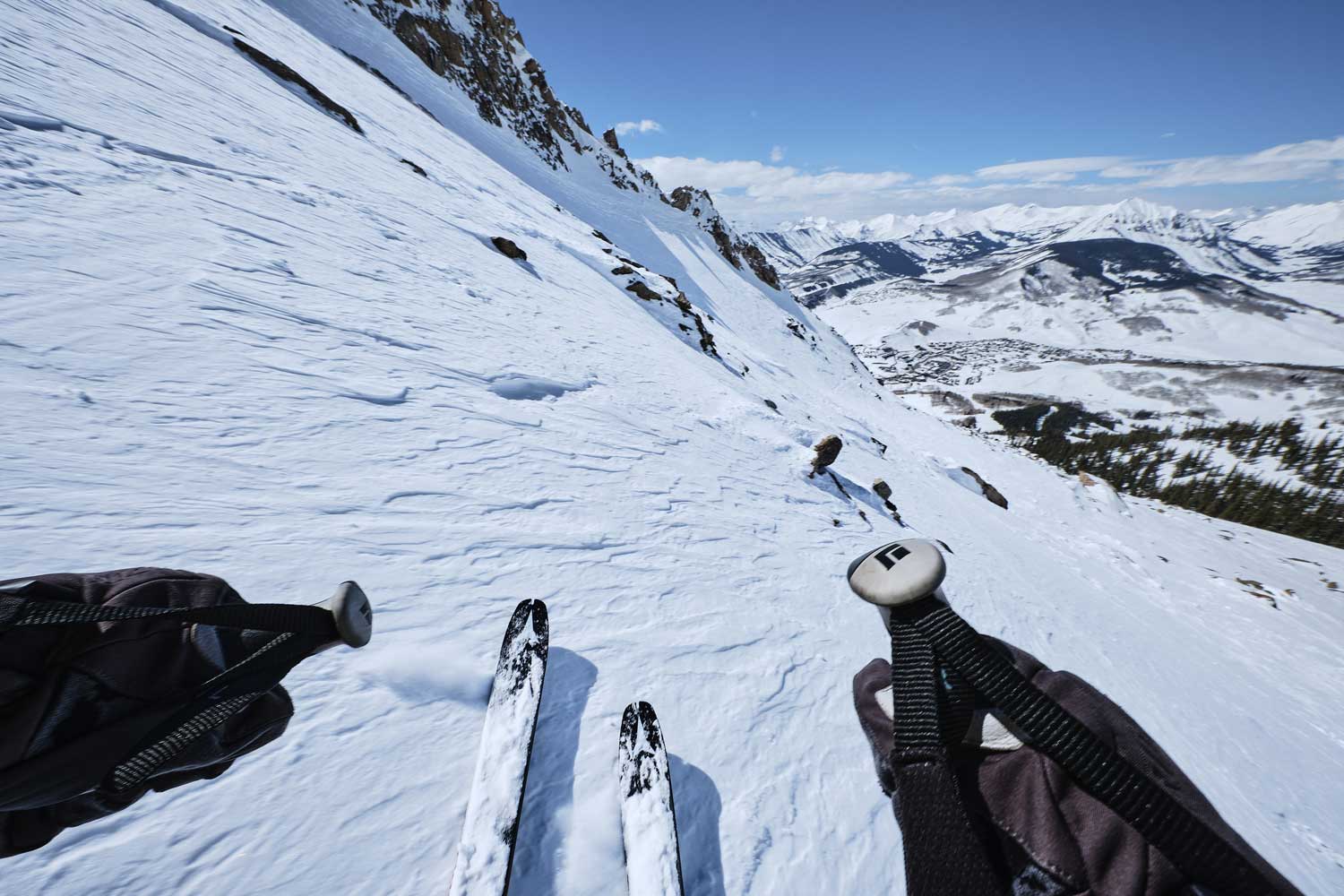 Point-of-view image of someone skiing down a snowy mountainside. The valley ahead is visible and the tips of the skis and hands holding ski poles enter the image from the bottom and bottom corners.