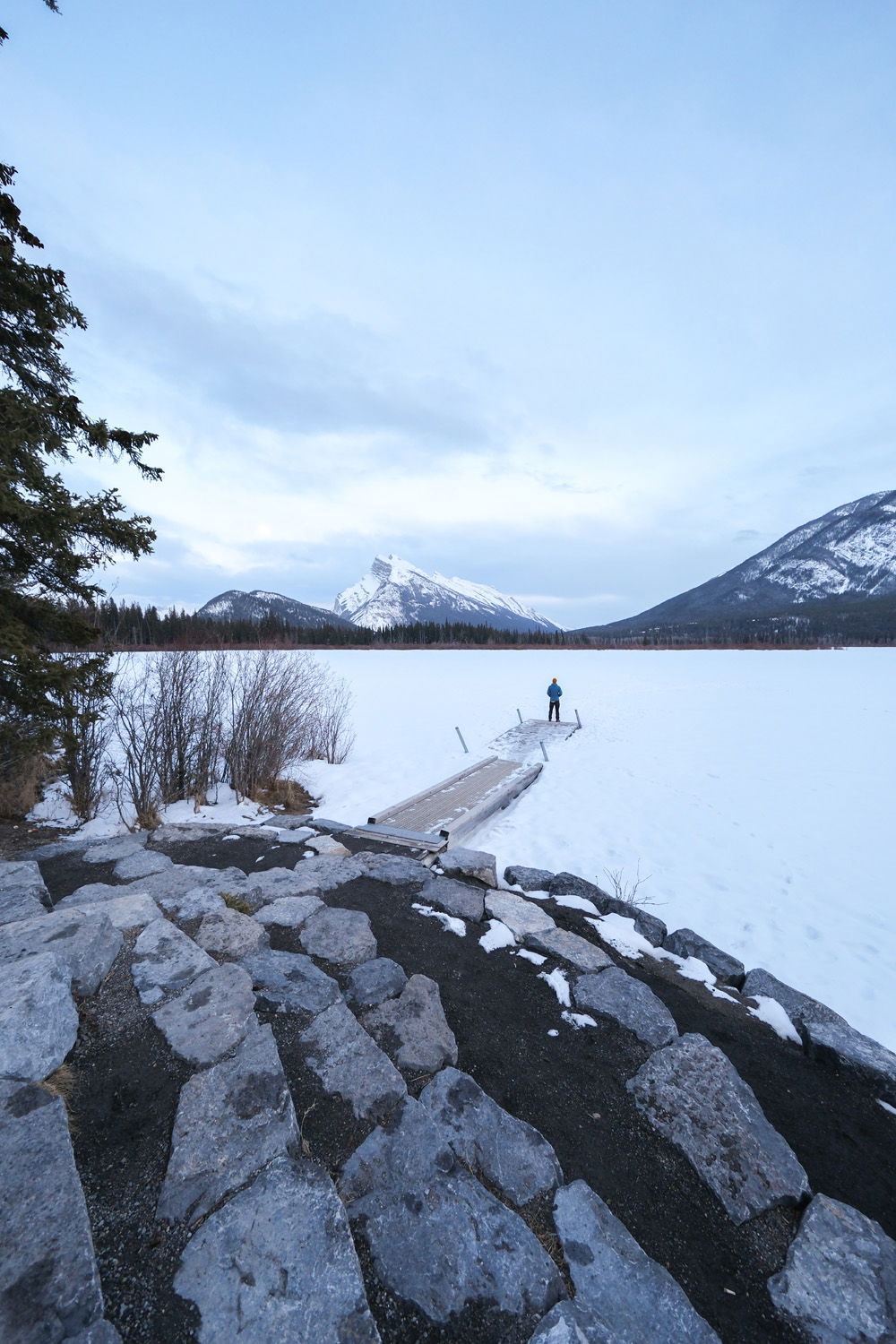 Wide shot of man standing at the end of a jetty out on a frozen lake with snowy mountains in the background