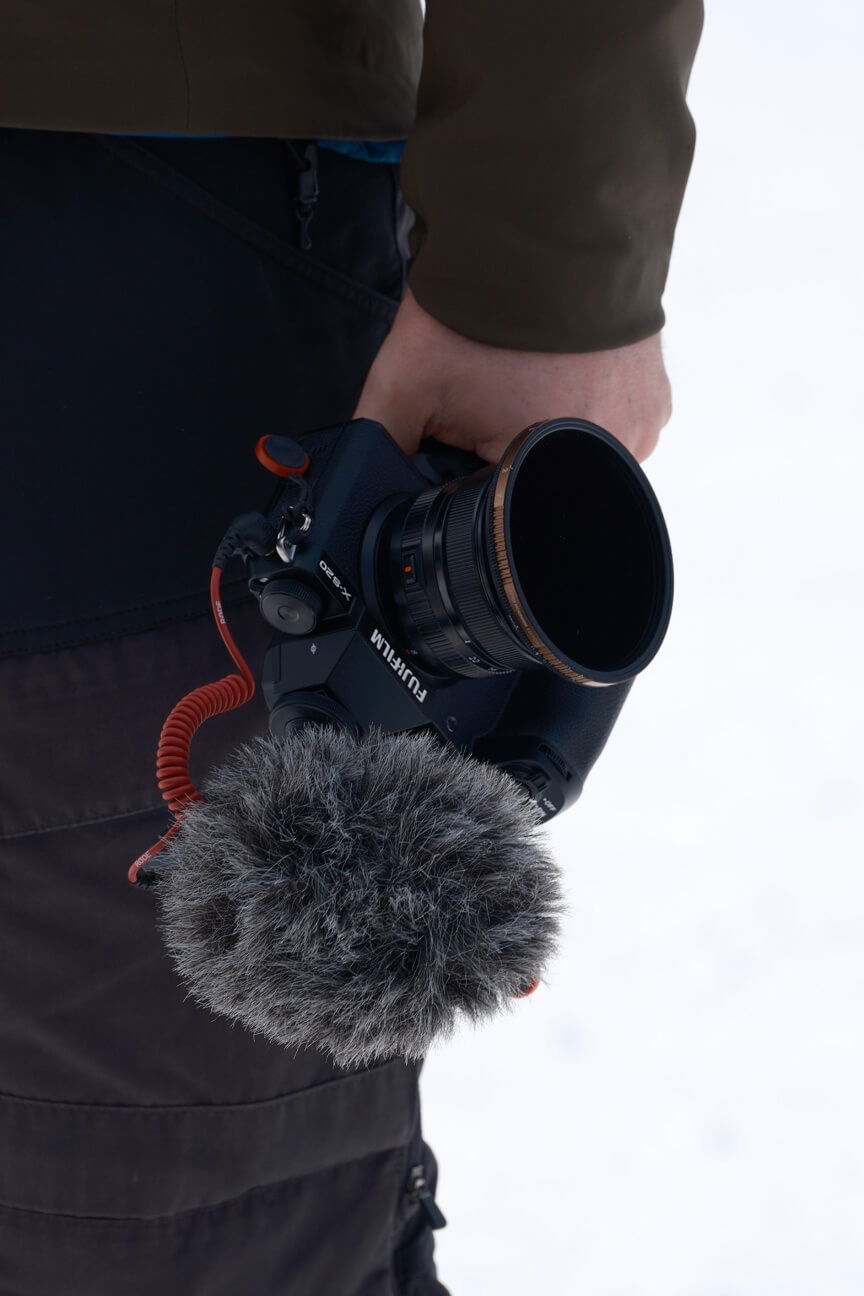Close up of person holding FUJIFILM X-S20 camera by their side. Camera is fitted with microphone and wind shield