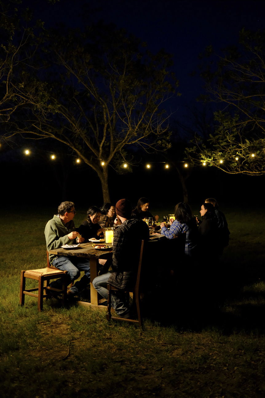 people eating outside on wooden table at night, illuminated by a string of lights