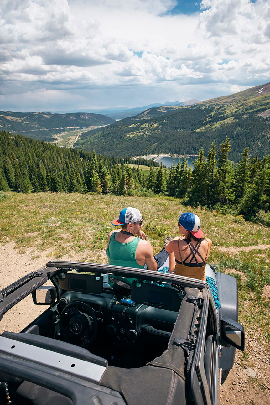 Man and woman sat on front of jeep in front of a valley landscape